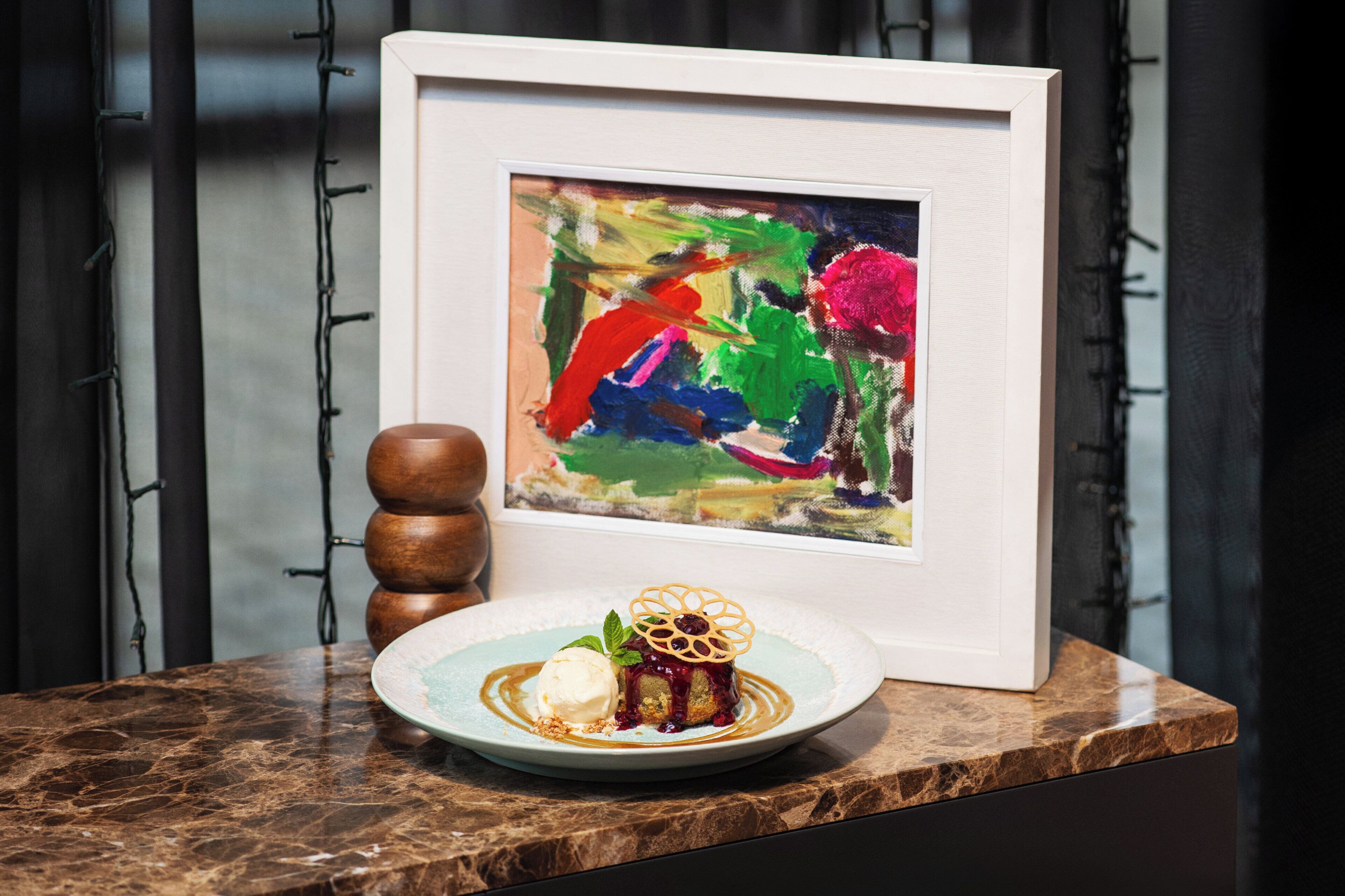 A dessert plate with a cake and ice cream on a marble countertop, a framed painting, and a wooden object.