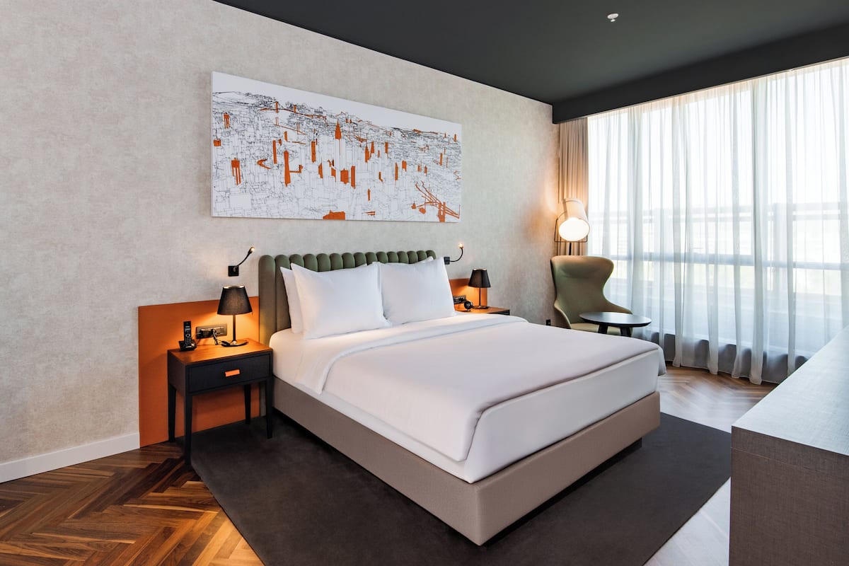Une chambre d’hôtel moderne avec un grand lit, une table de chevet avec une lampe, une chaise et une grande fenêtre avec des rideaux légers.