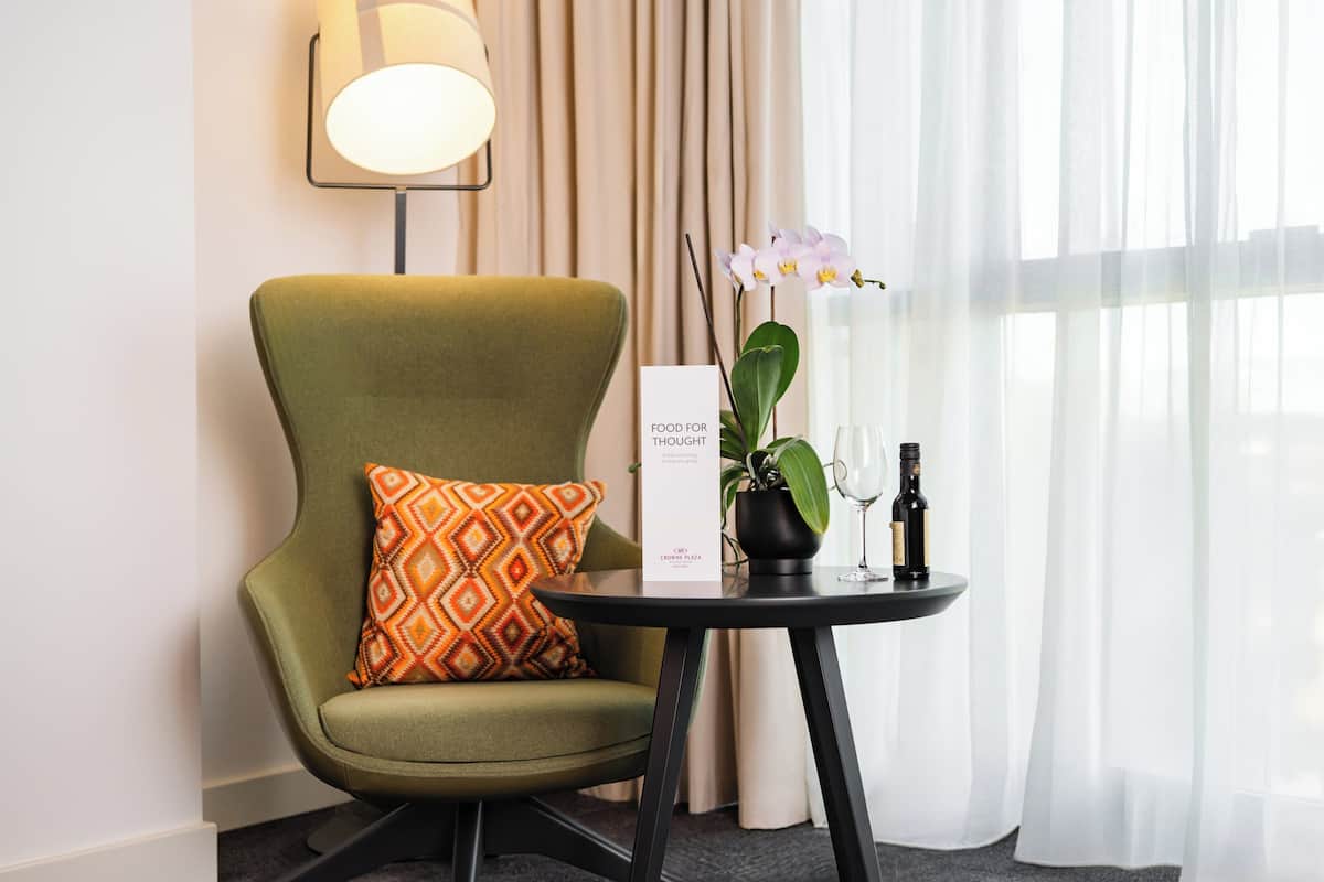 Une chambre d’hôtel avec un fauteuil vert, une petite table sur laquelle se trouvent une bouteille de vin et un verre, une orchidée en pot, et une pancarte indiquant le nom et l’emplacement de la chambre.