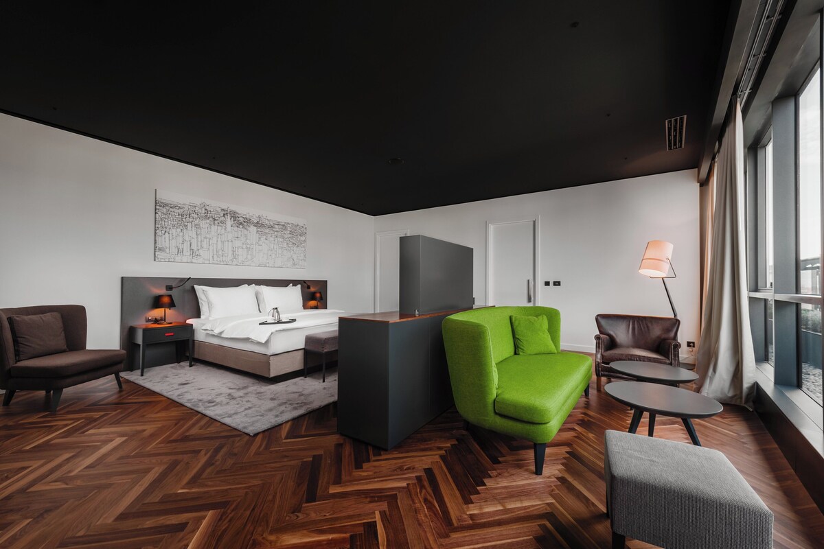 Une chambre d’hôtel moderne avec un lit, un fauteuil vert, un fauteuil marron, une petite table et une lampe.