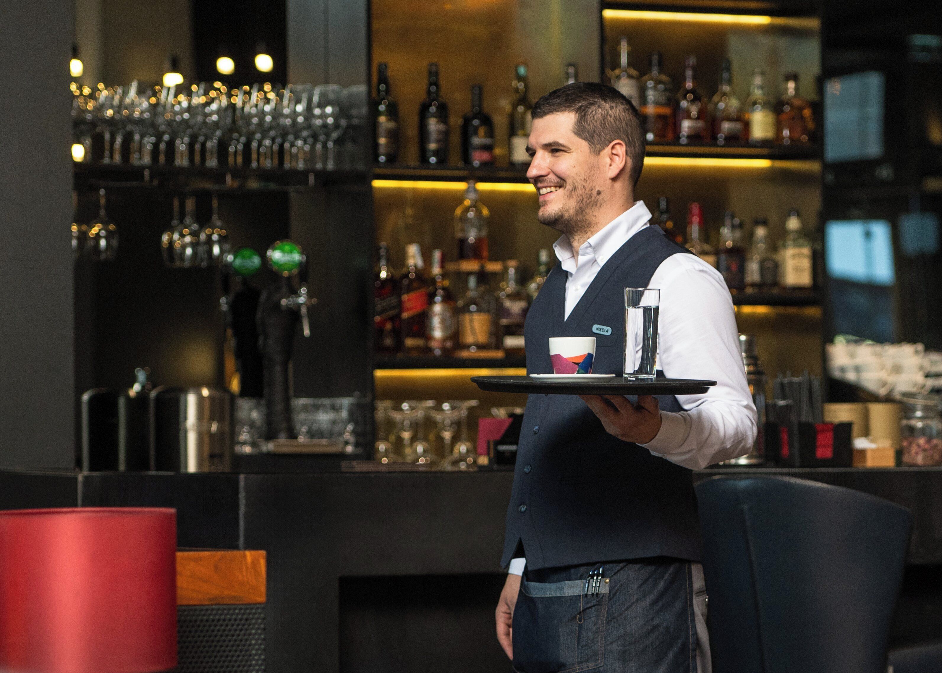 Una zona de bar con estantes llenos de botellas y vasos, una barra y un cubo de basura rojo.