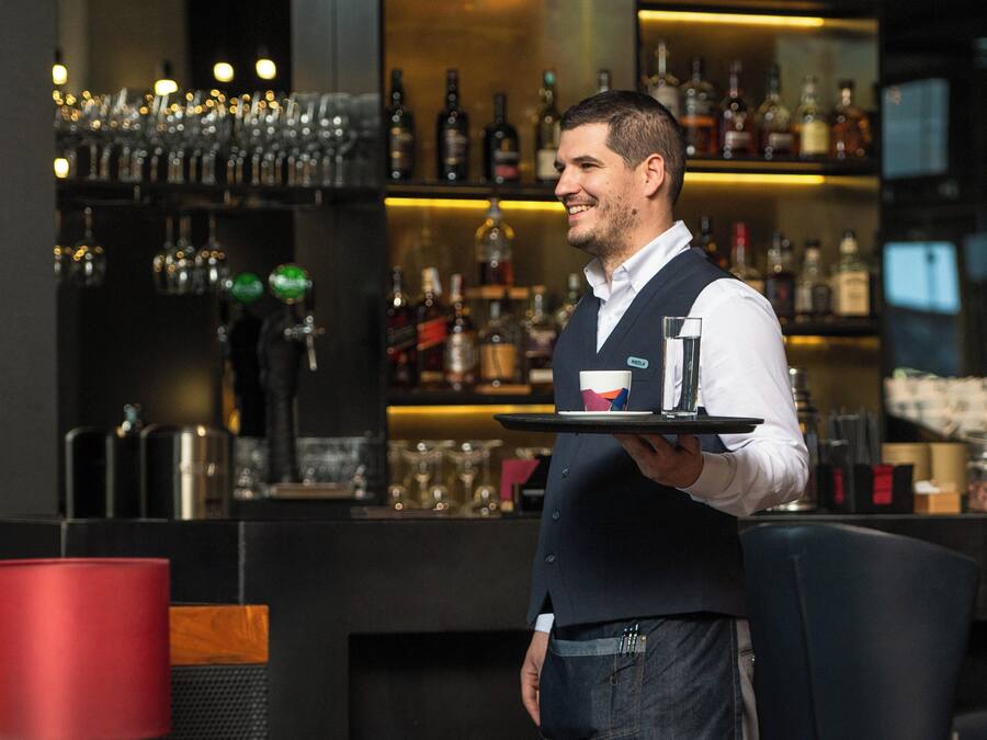 Una zona de bar con estantes llenos de botellas y vasos, una barra y un cubo de basura rojo.