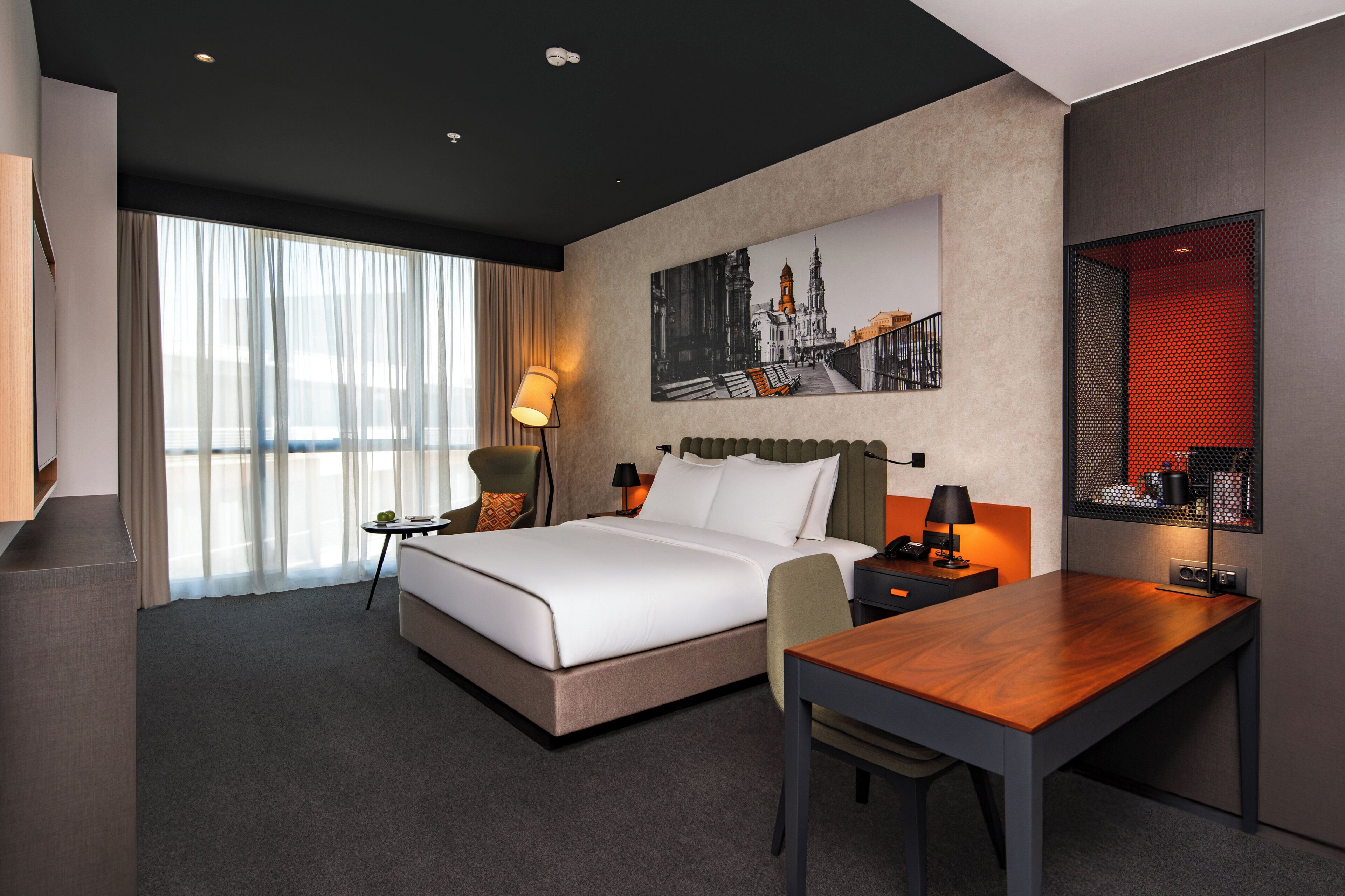 A modern hotel room with a large bed, a desk, a chair, a lamp, and a large window with sheer curtains.