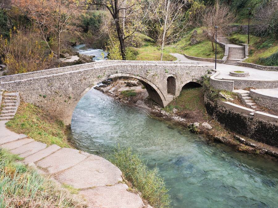 A stone bridge over a clear stream, surrounded by trees and a stone pathway.