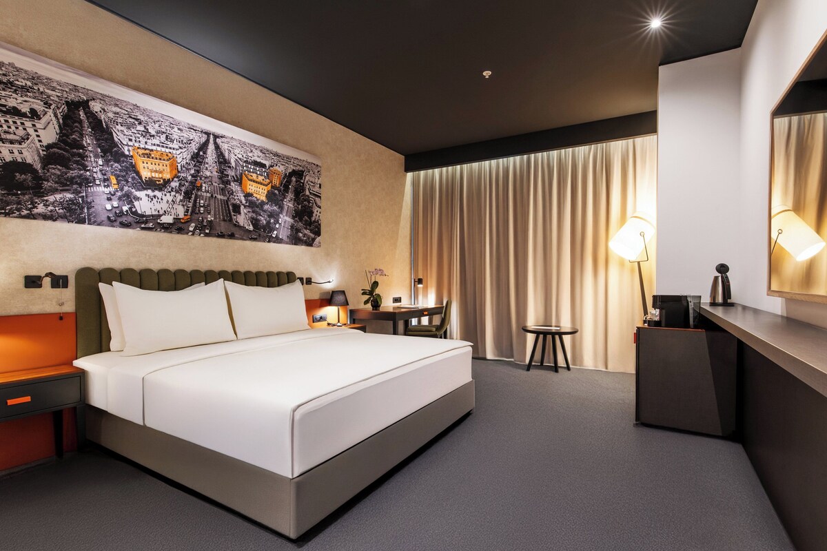 Une chambre d’hôtel moderne, dotée d’un grand lit, d’un bureau, d’une chaise, d’une lampe et d’un grand tableau au mur.