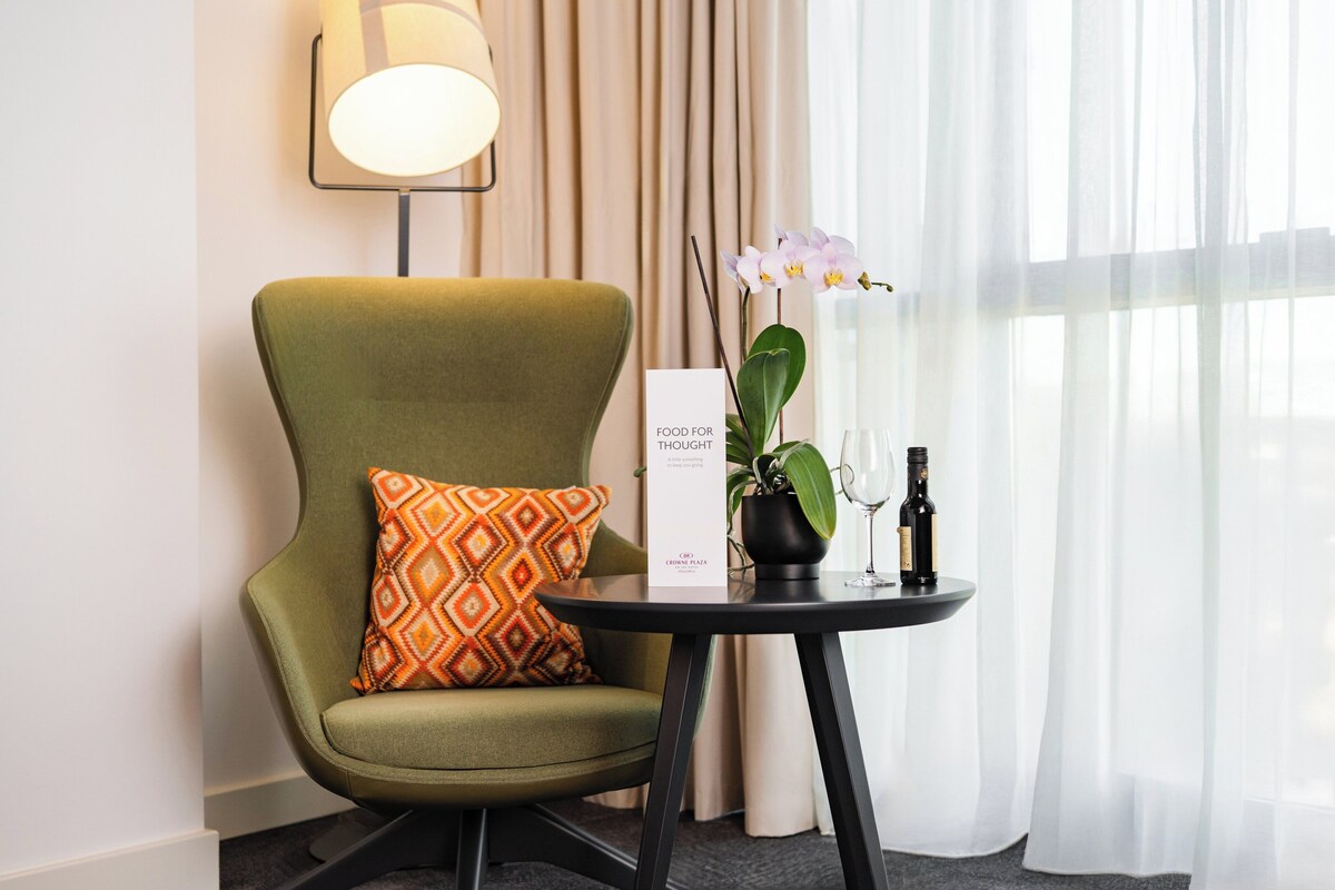 Un fauteuil vert agrémenté d’un coussin à motifs, une petite table avec une bouteille de vin et un verre, une orchidée en pot et une pancarte indiquant « À MEDITER », le tout dans un décor d’hôtel.