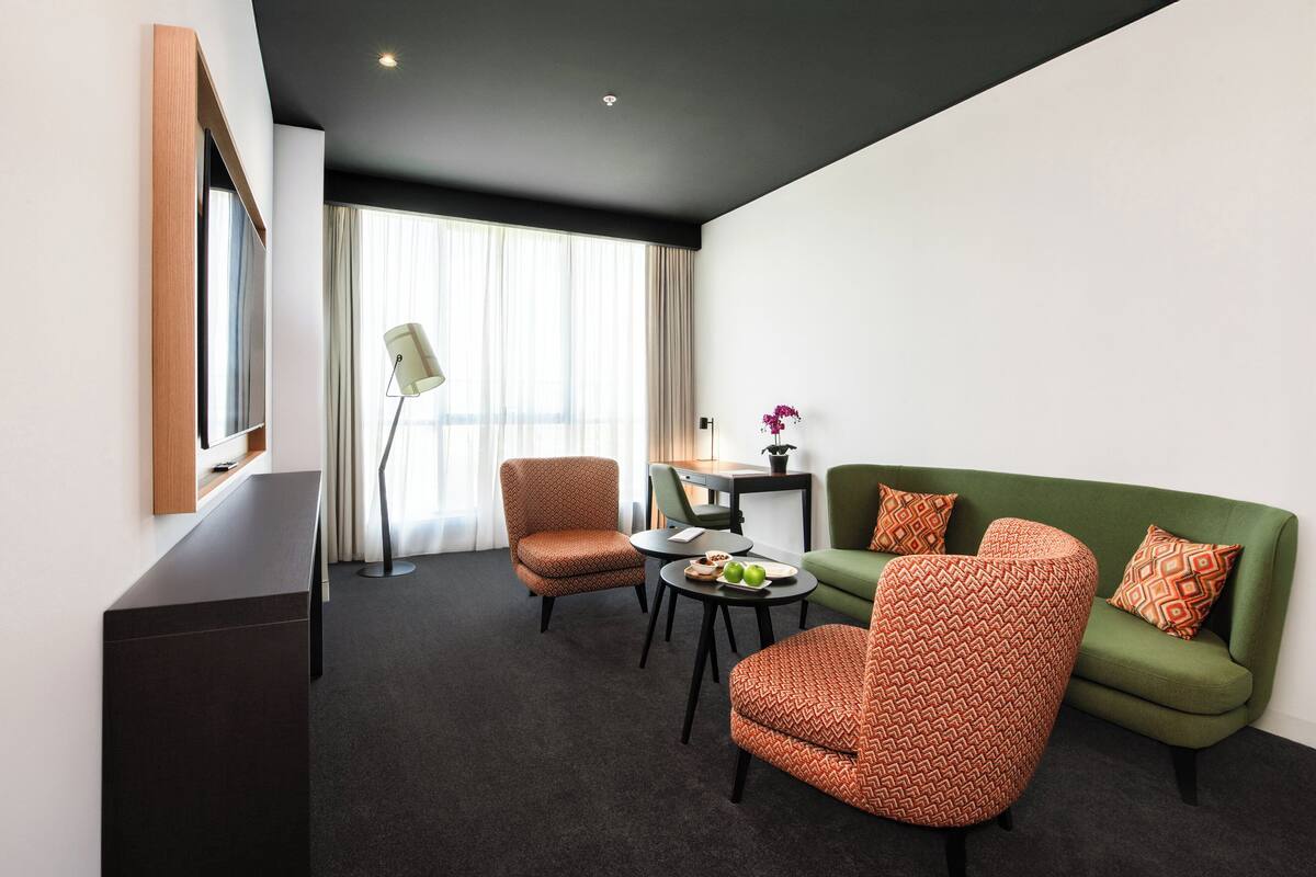 Une chambre d’hôtel moderne avec un plafond sombre, un canapé vert, deux fauteuils à motifs, une petite table ronde avec une lampe et une grande fenêtre avec des rideaux.