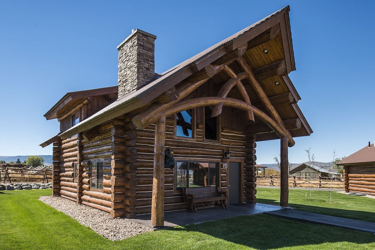 A log cabin with a stone chimney, large windows, and a porch with a bench.