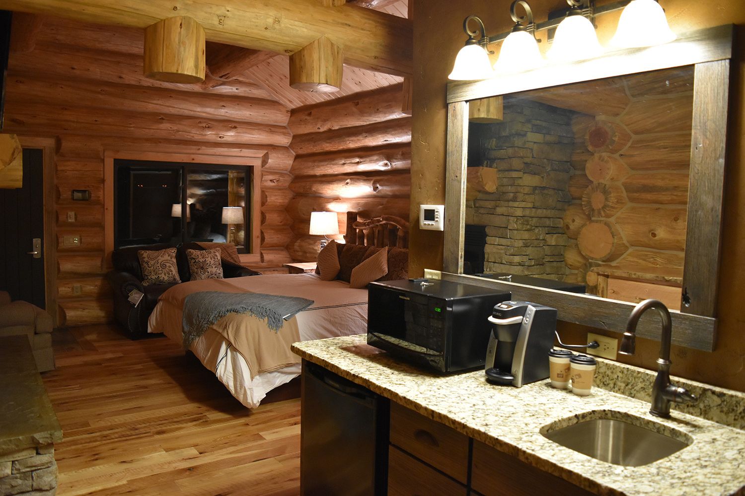 Una habitación tipo cabaña de madera con una cama, cafetera, lavamanos y chimenea de piedra.