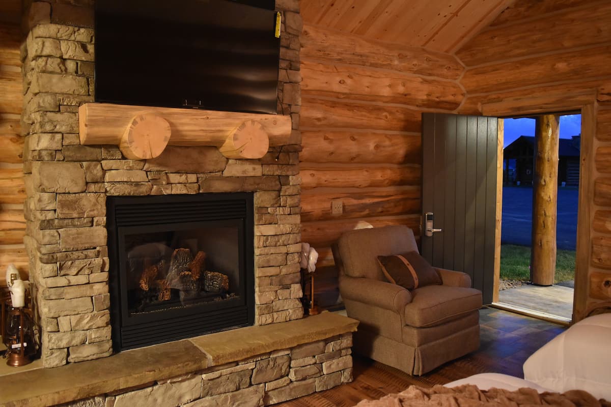 A cozy log cabin interior with a stone fireplace, a comfortable armchair, and a view of the outside.