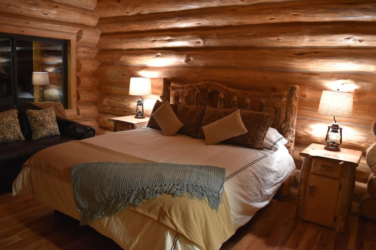 A log cabin bedroom with a large bed, two bedside tables with lamps, a wooden headboard, and a window with a view.