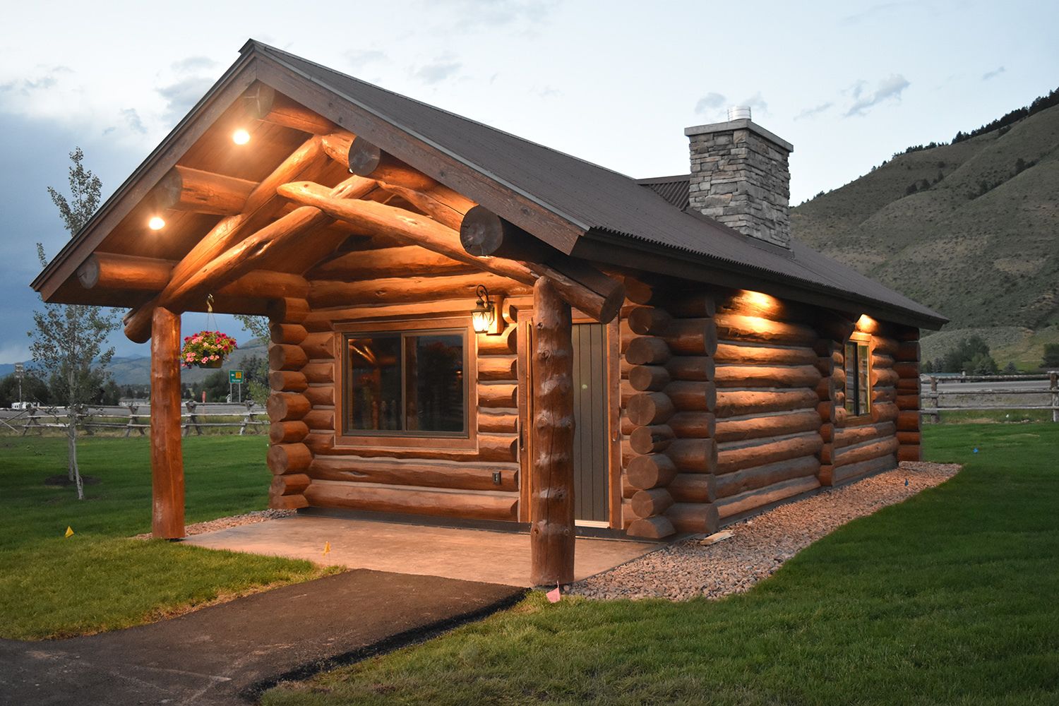 A log cabin with a stone chimney, a porch with hanging flower baskets, and a well-lit entrance.