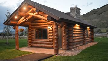 A log cabin with a stone chimney, a porch with hanging flower baskets, and a well-lit entrance.
