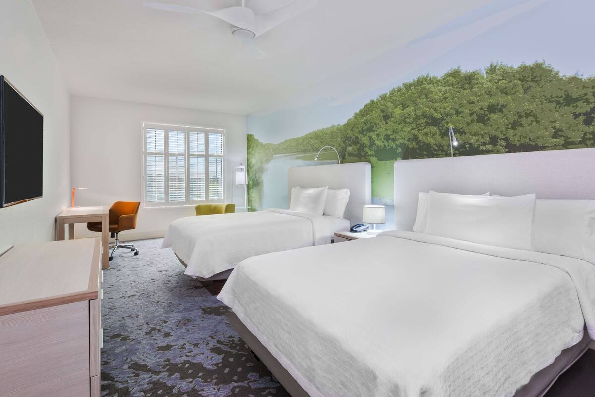 A hotel room with two beds, a desk, a chair, and a large window.