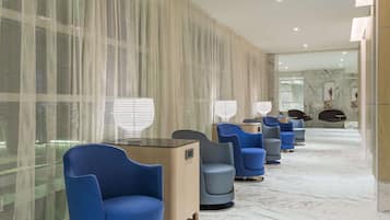 A modern hotel lobby with a row of blue chairs, a reception desk, and a glass partition with a mesh design.