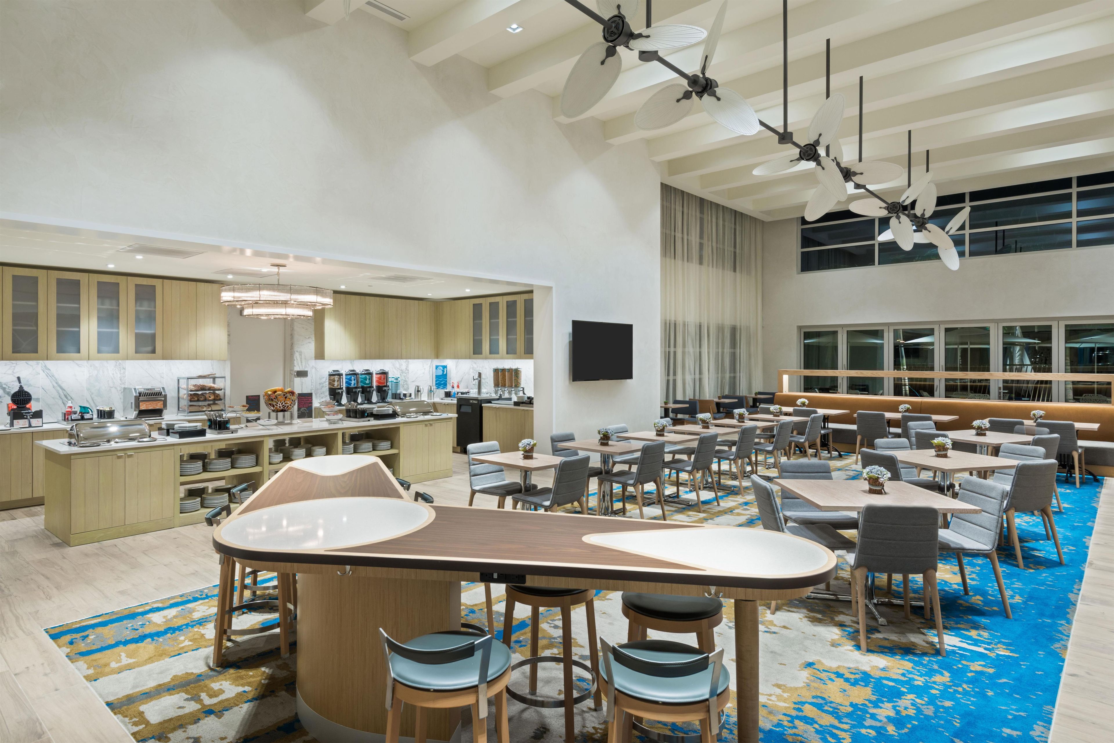 A spacious dining area with multiple tables and chairs, a kitchen in the background, and a large window providing natural light.