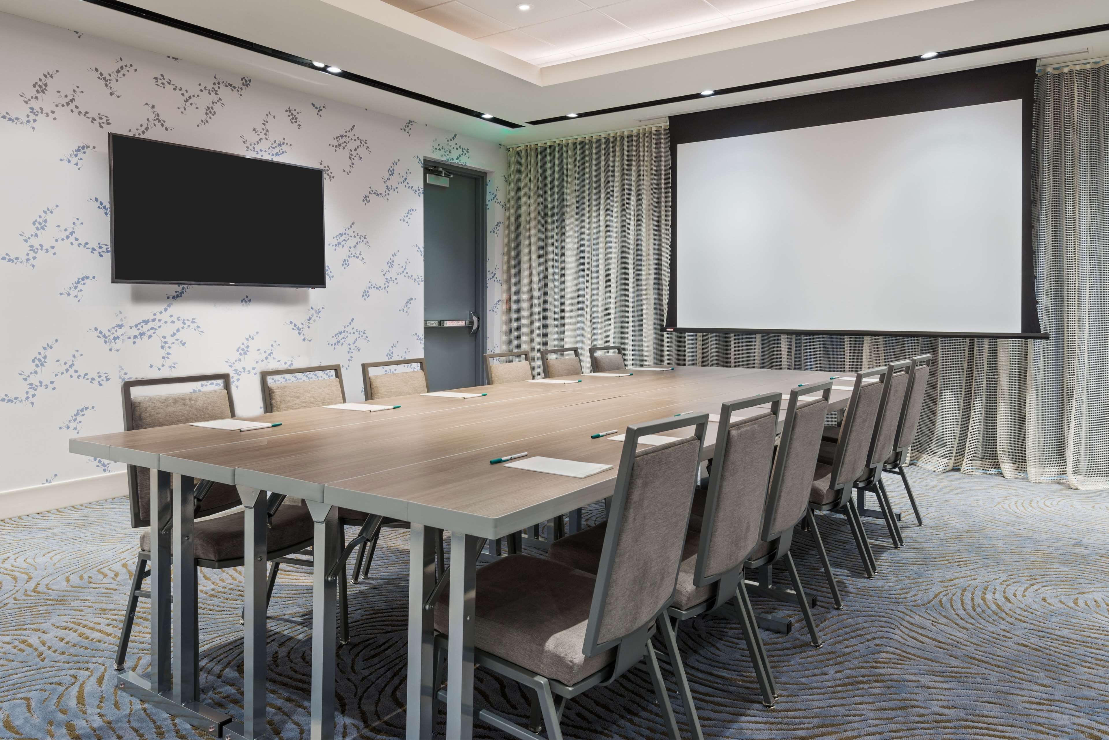 Ein Konferenzraum mit langem Tisch, Stühlen, einer Projektionsleinwand und einem Fernseher.