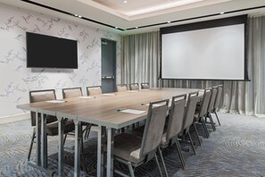 Ein Konferenzraum mit langem Tisch, Stühlen, einem Flachbildfernseher, einer Projektionsleinwand und ornamentalem Wandpapier.