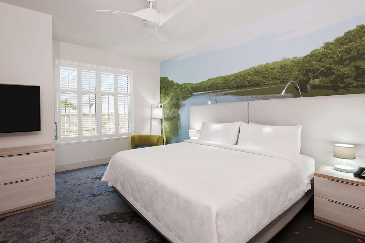 A hotel room with a bed, a TV, a desk, a chair, and a window with blinds.