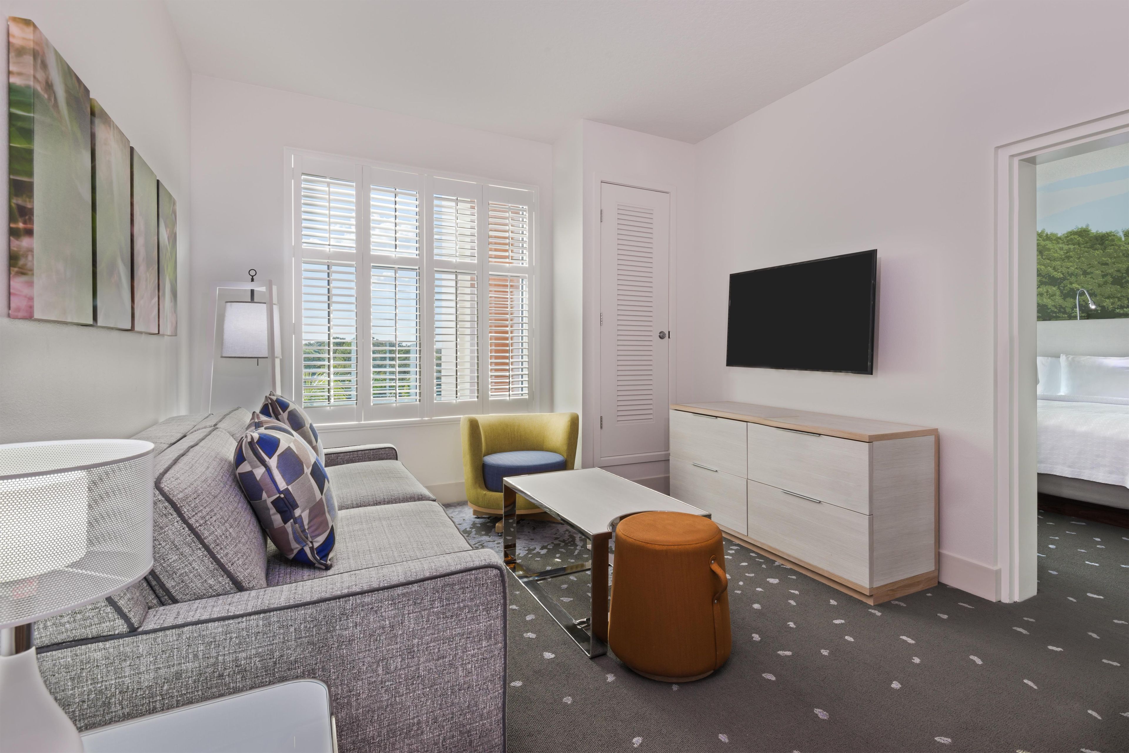 A modern, well-lit room with a sofa, armchair, and a small table. A television is mounted on the wall, and there is a door leading to another area.