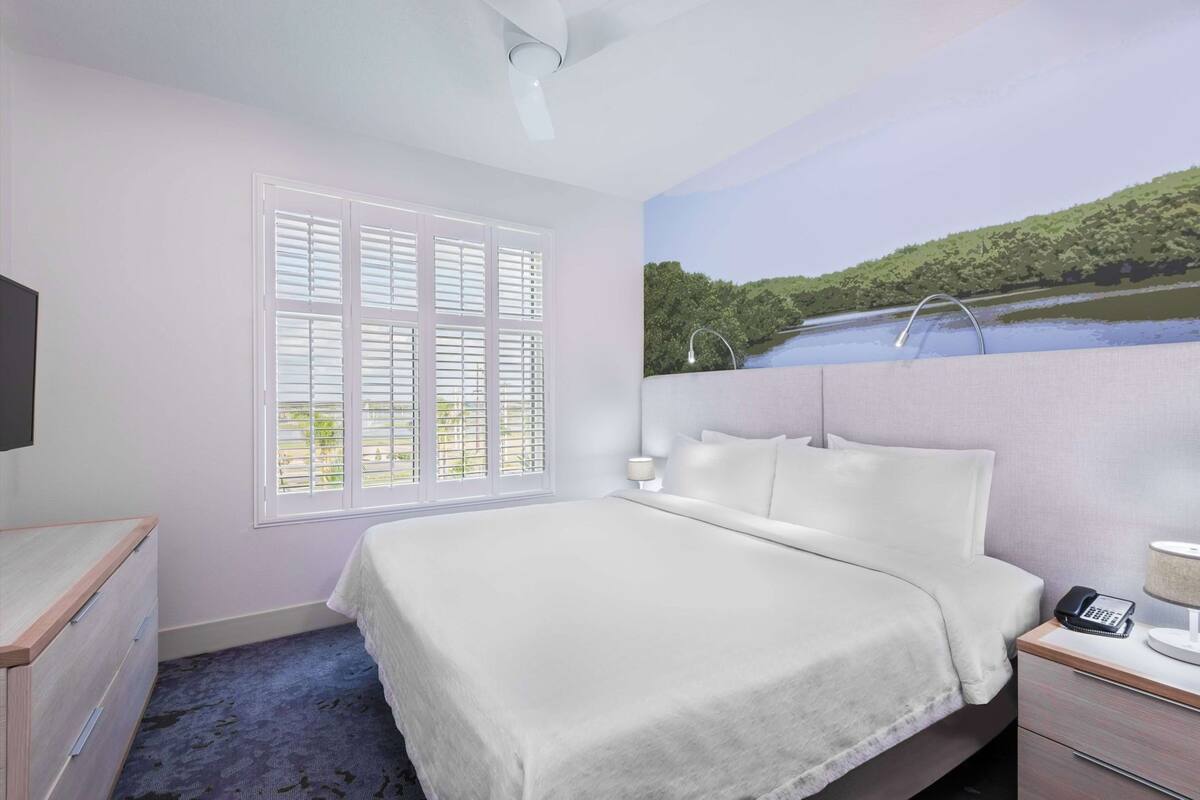 A hotel room with a bed, a desk, a chair, a lamp, and a painting of a landscape.
