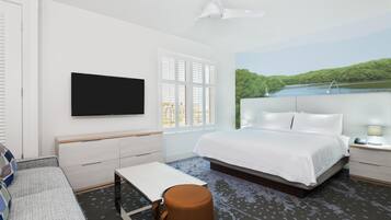 A hotel room with a bed, a TV, a desk, a chair, a window with blinds, and a painting of a lake.