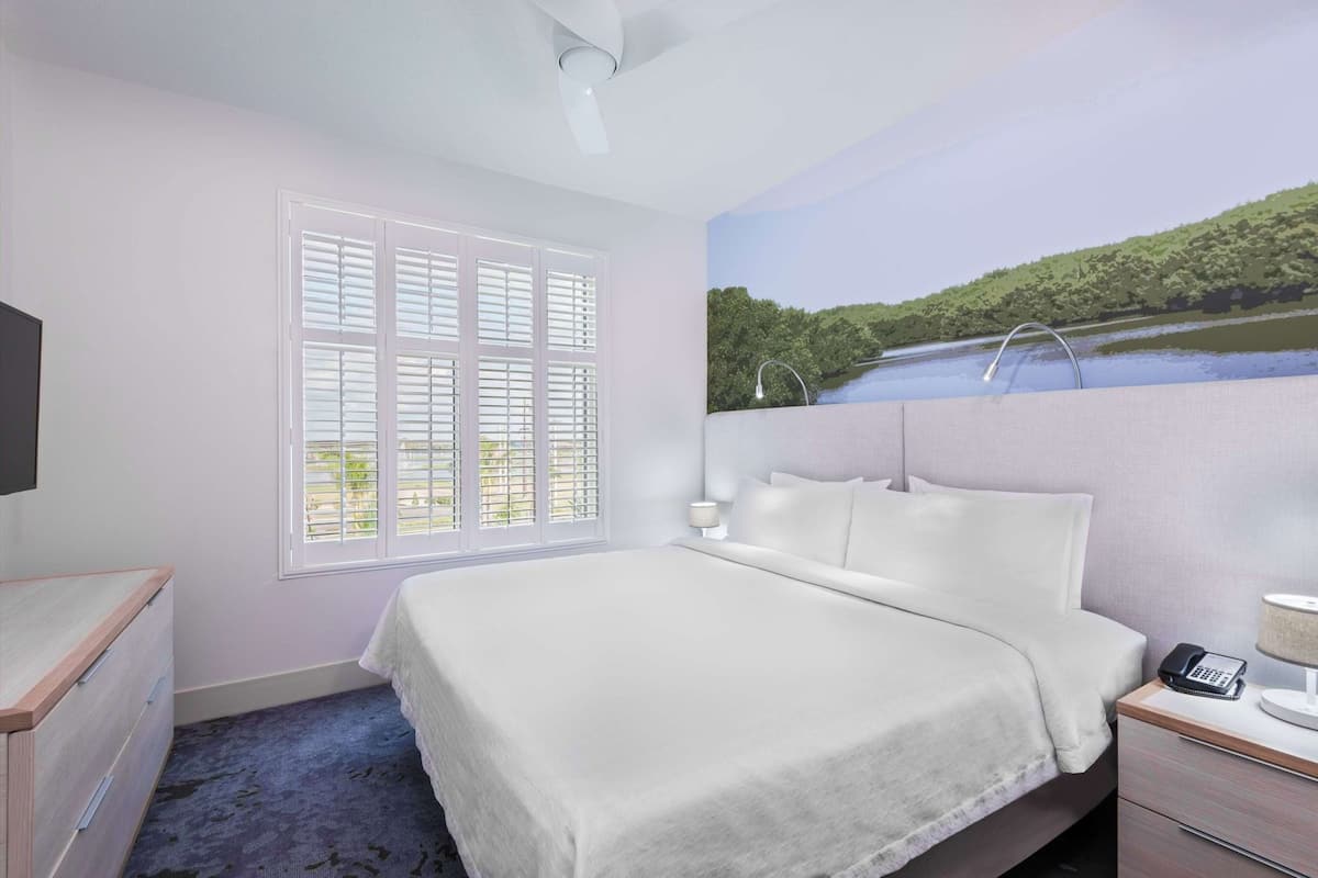 A hotel room with a bed, a desk, a chair, a lamp, and a painting of a landscape.