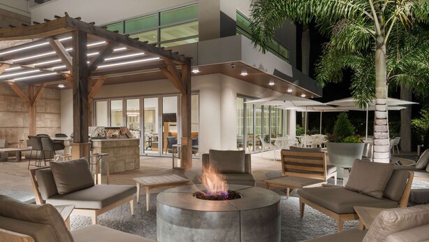 A modern outdoor lounge area with a circular fire pit surrounded by comfortable seating, a pergola overhead, and a view of the interior space. - Homewood Suites by Hilton Sarasota Lakewood Ranch (Sarasota)