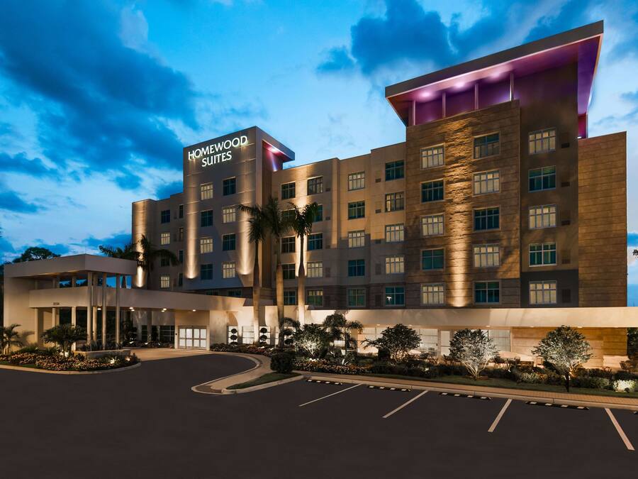 Um hotel moderno com uma placa grande de "Homewood Suites", vários andares e uma área de entrada bem iluminada com estacionamento.