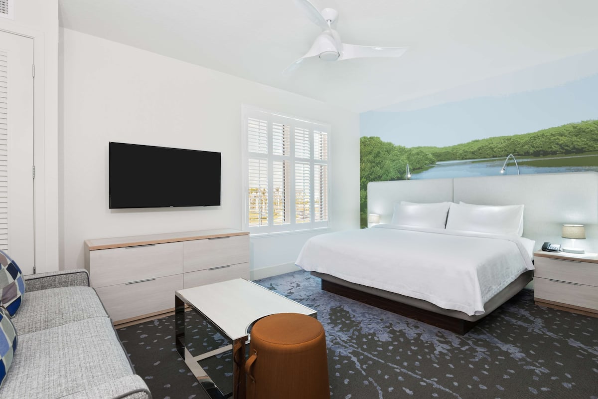 A hotel room with a bed, a TV mounted on the wall, a window with blinds, a ceiling fan, and a painting of a lake.