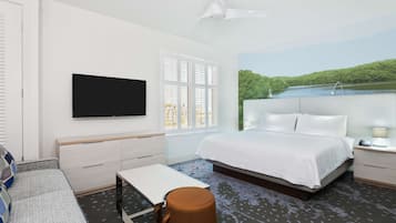 A hotel room with a bed, a TV mounted on the wall, a window with blinds, a ceiling fan, and a painting of a lake.