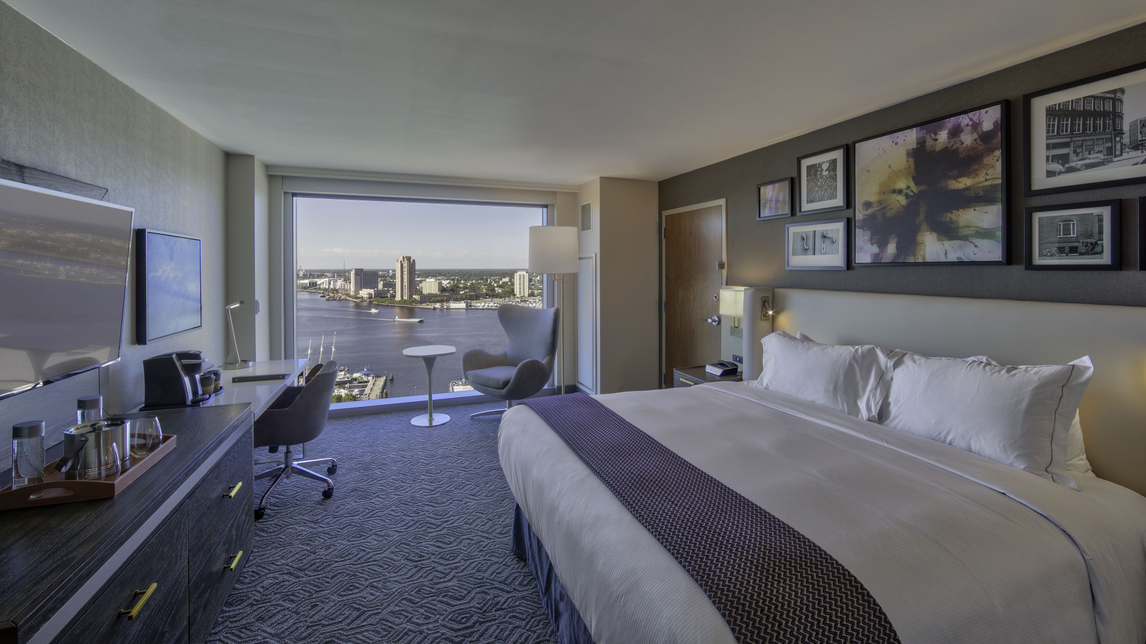 A hotel room with a large bed, a desk with a chair, a TV, and a view of the city through a large window.