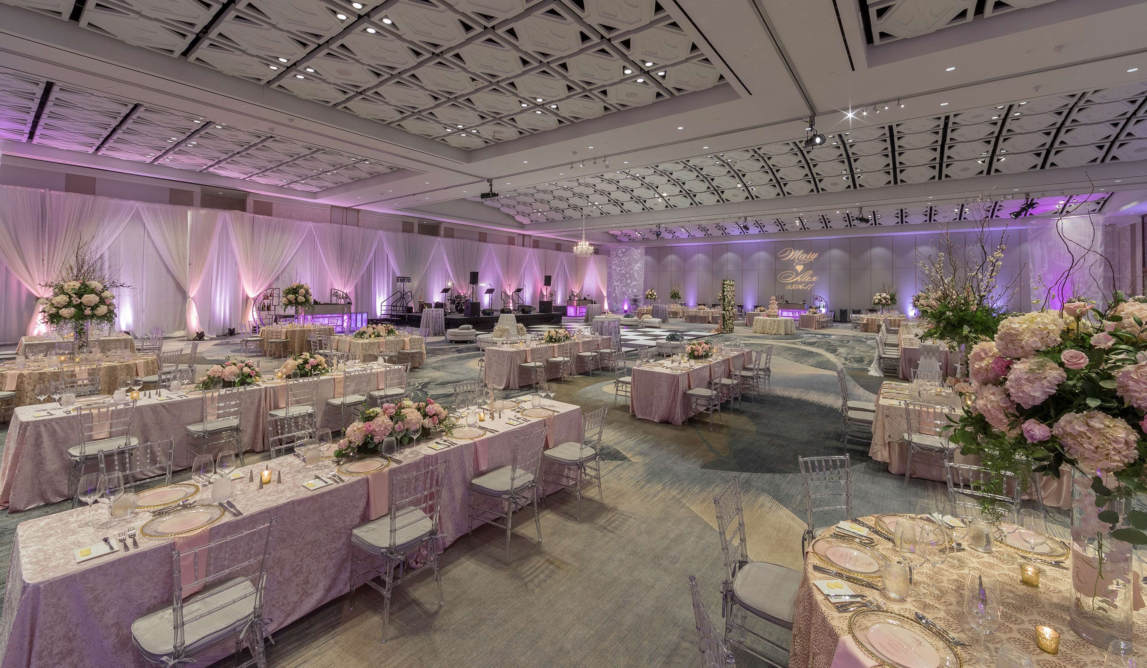 A spacious banquet hall with round tables, clear chairs, and elegant floral centerpieces.