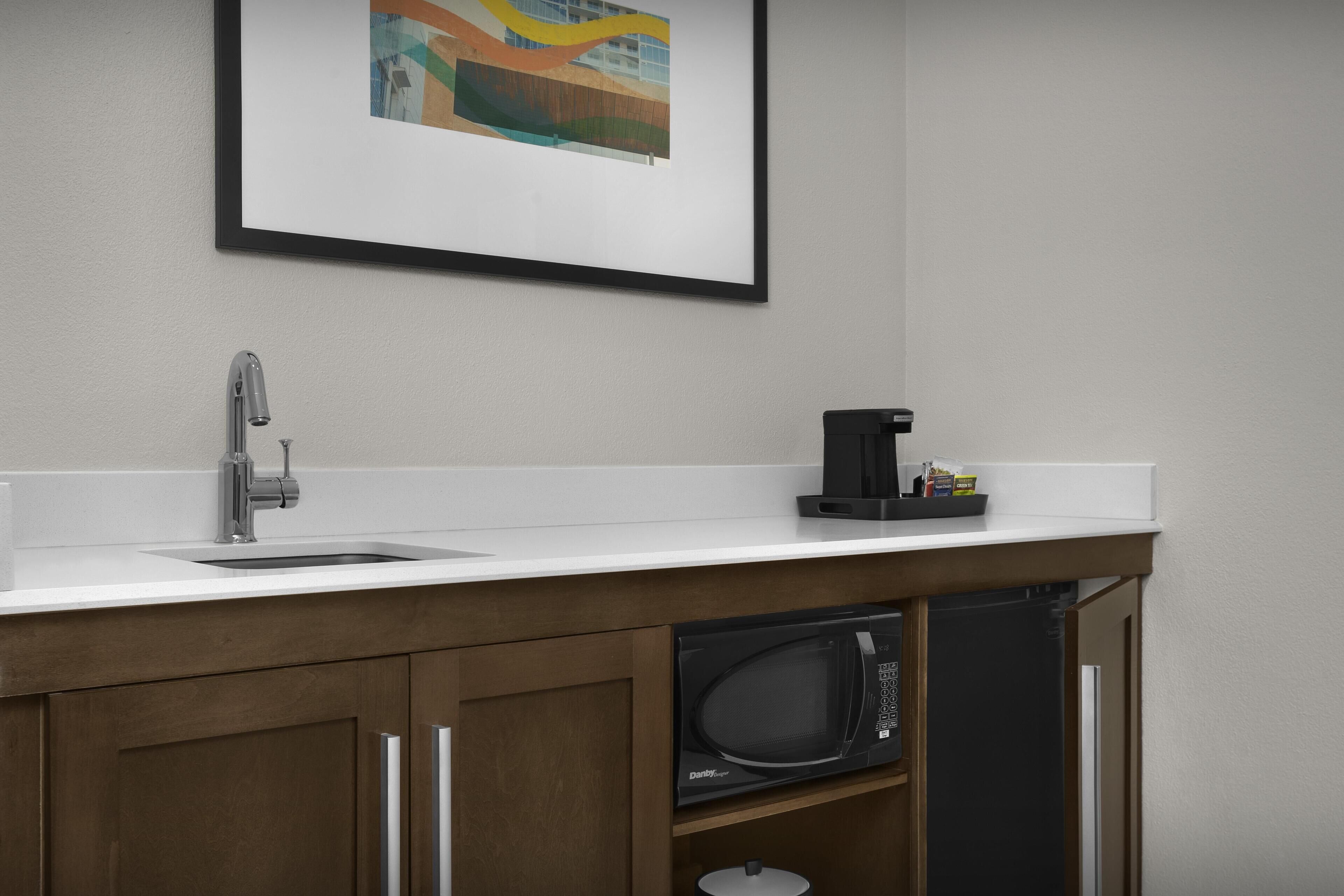 Cozinha com bancada branca, torneira de aço inoxidável, cafeteira preta, micro-ondas e um quadro na parede.