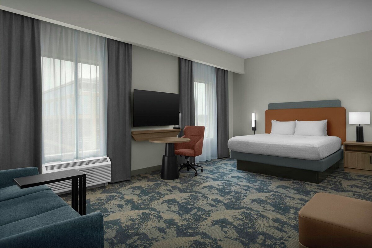 A hotel room with a bed, a desk with a chair, a TV, and a window with curtains.