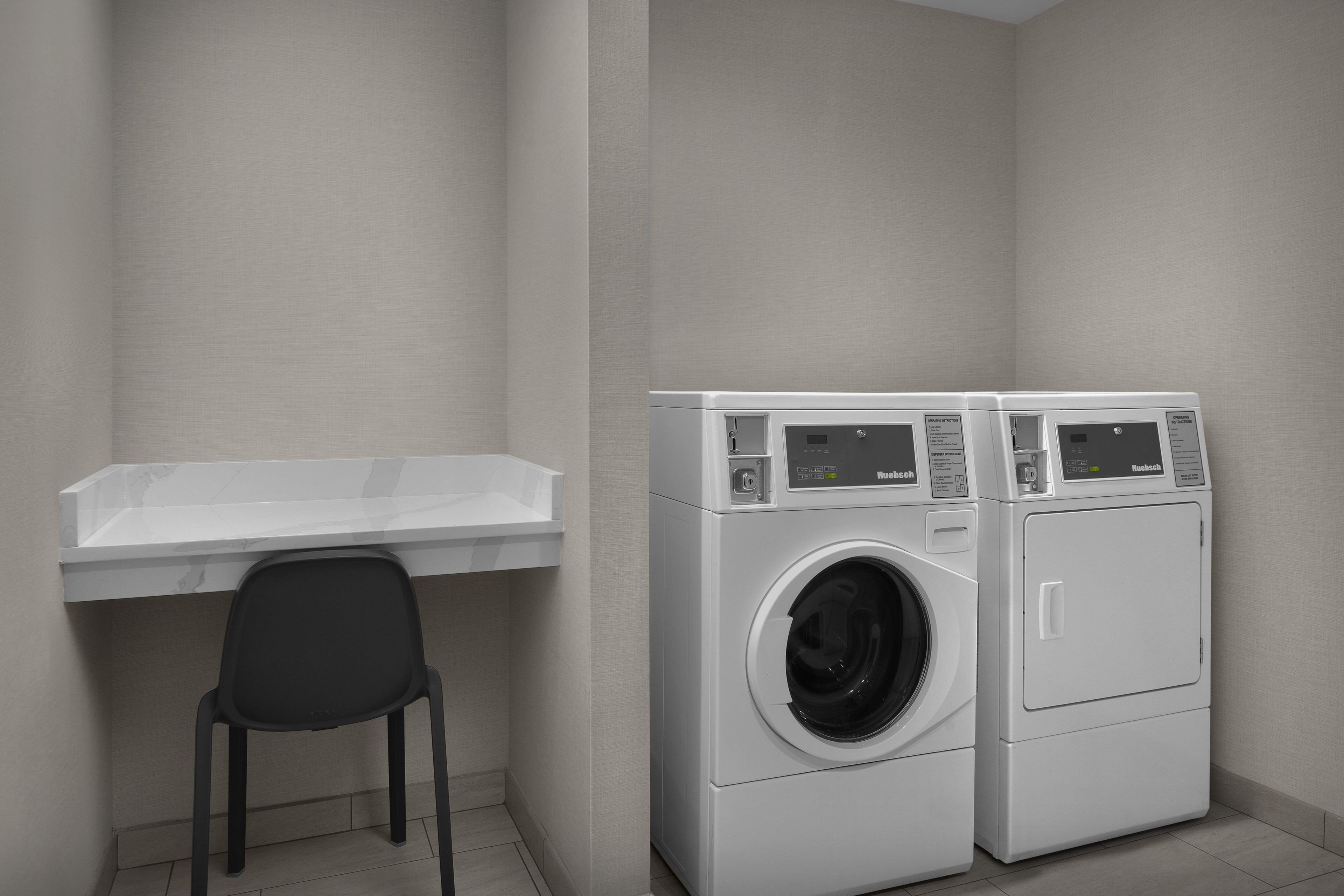 A laundry room with two front-loading washing machines, a small chair, and a white countertop.