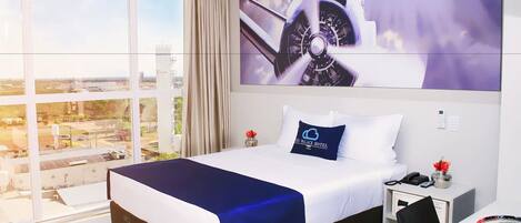 Habitación de hotel con una cama grande, un escritorio con una computadora portátil, una ventana con vista a la ciudad y un televisor montado en la pared.