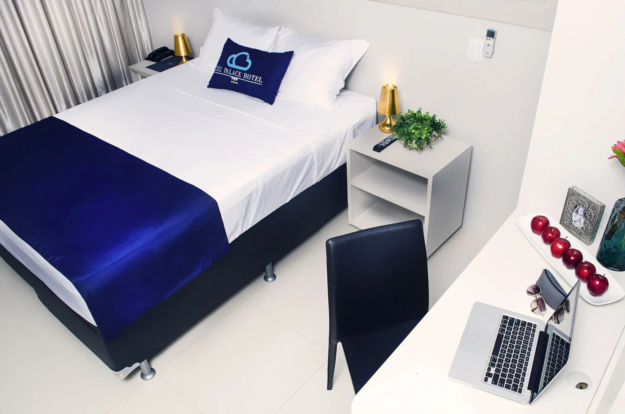 A hotel room with a bed, a desk with a laptop, a chair, a bedside table with a lamp, and a nightstand with a phone.