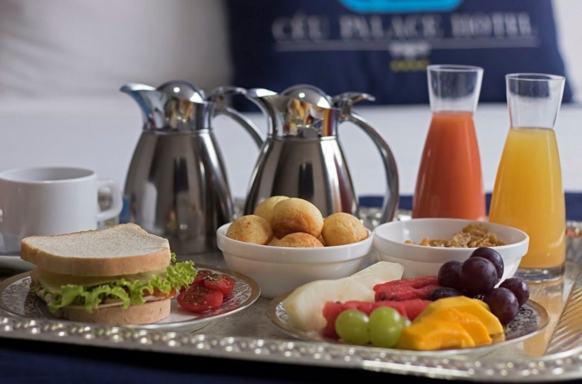 Bandeja de desayuno con sándwich, croquetas, fruta, cereal y jugo.