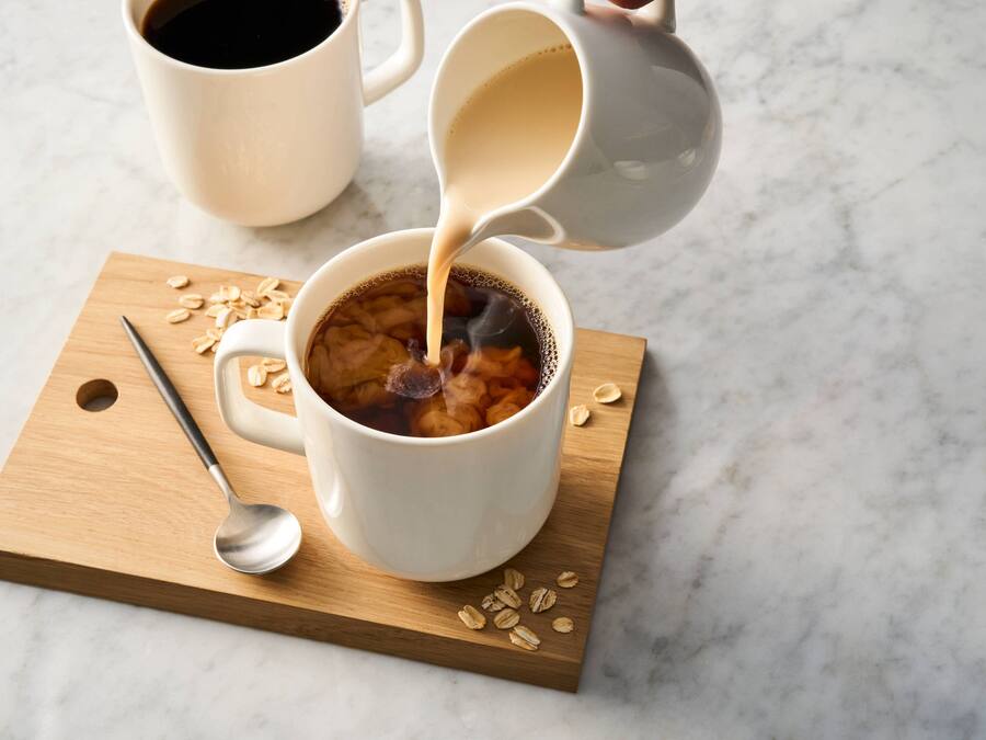 A white mug on a wooden board with oats and a spoon, and a white mug being filled with a dark liquid.