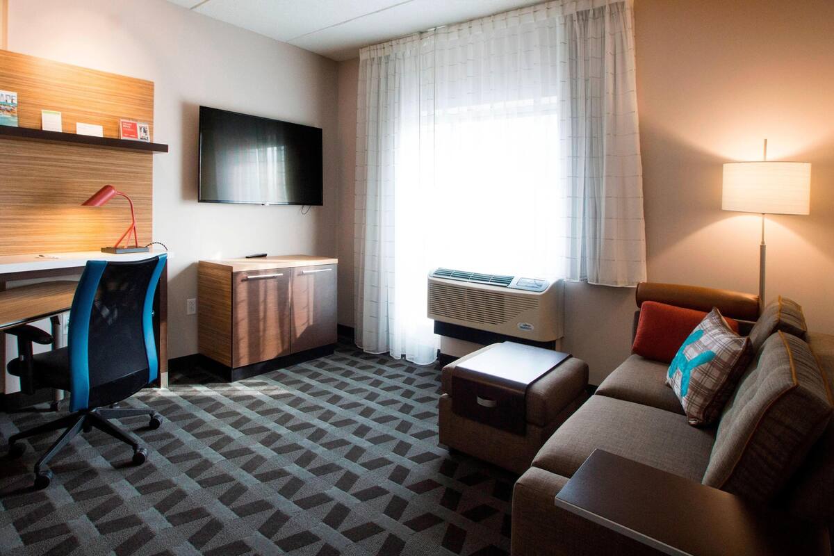 A hotel room with a desk, chair, sofa, TV, air conditioning unit, and a window with curtains.