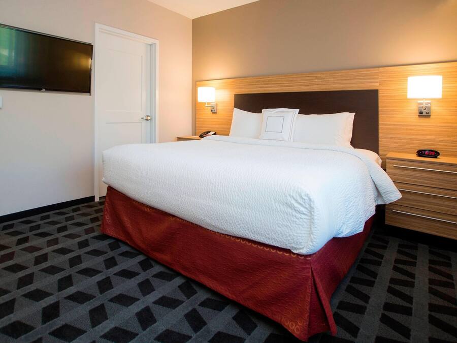 A hotel room with a bed, bedside tables, a TV, and a wall-mounted light.