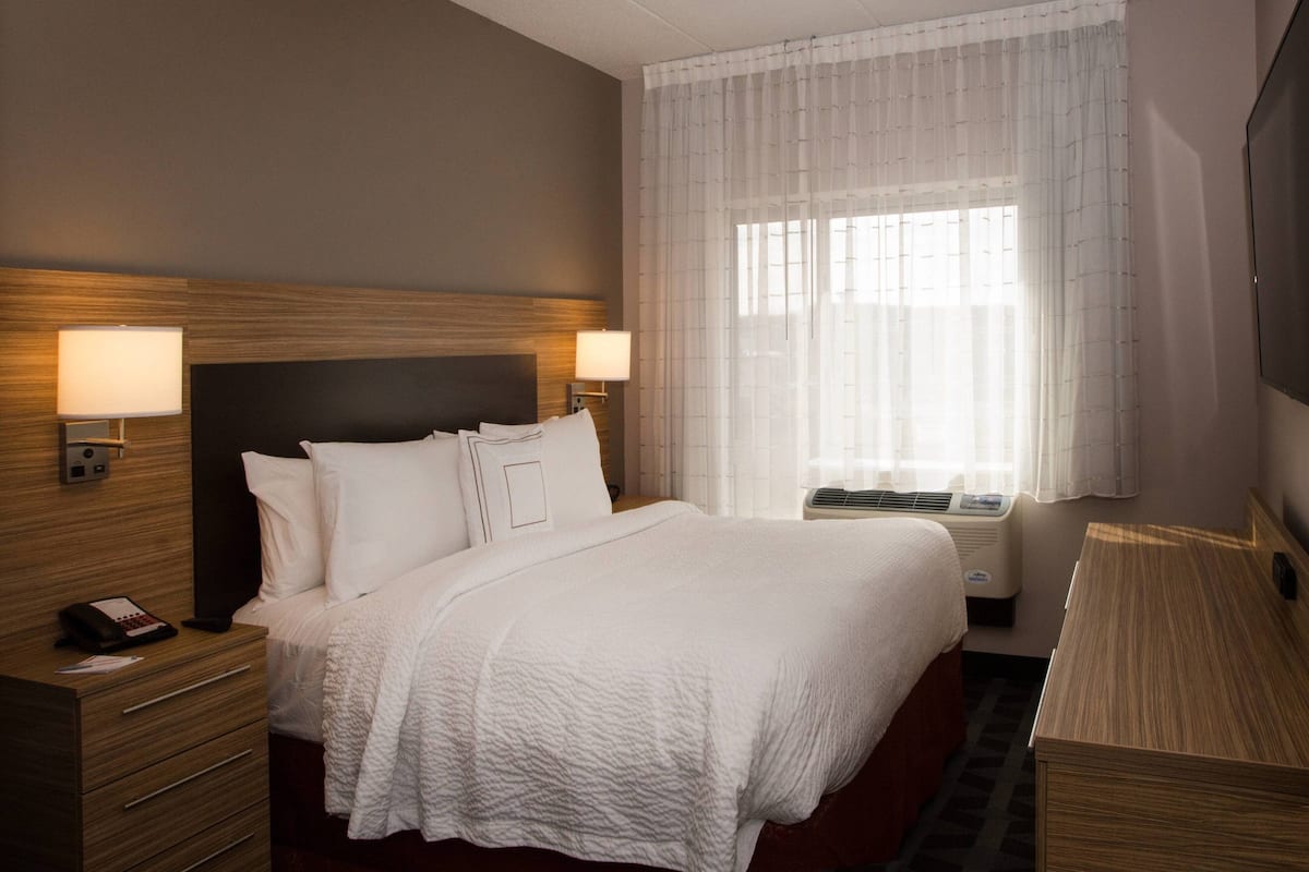A hotel room with a large bed, two bedside lamps, a nightstand with a phone, and a window with sheer curtains.