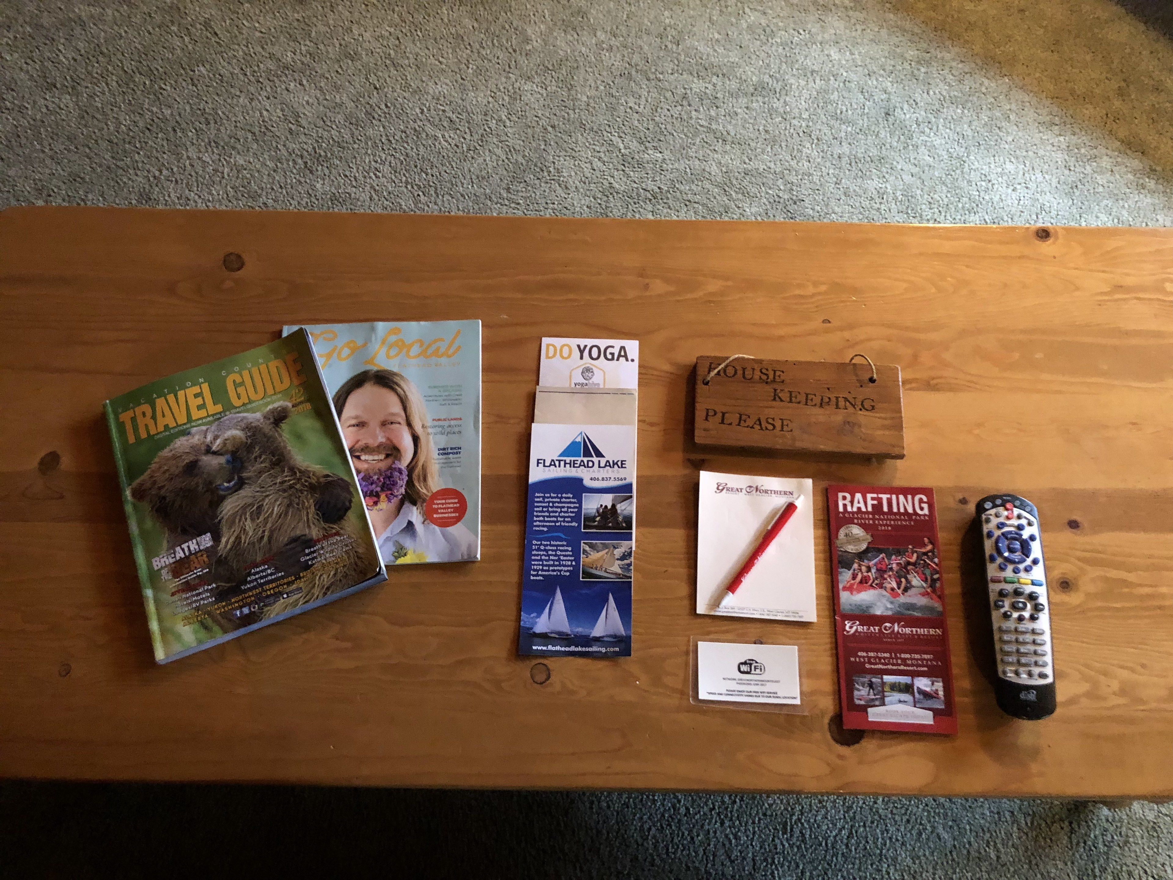 A wooden table with various items including a travel guide, a yoga pamphlet, a Wi-Fi sign, a remote control, and a sign that says "HOUSE KEEPING PLEASE".