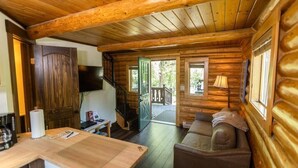 A log cabin interior with a wooden dining table, a sofa, a television, and a staircase leading to an outdoor deck.