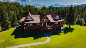 A large, multi-story wooden lodge with a wrap-around porch, set in a forested area with a clear view of distant mountains.