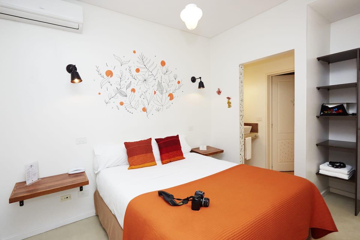 A neatly arranged hotel room with a bed, a bedside table with a lamp, a wall-mounted air conditioner, and a shelf with a camera.