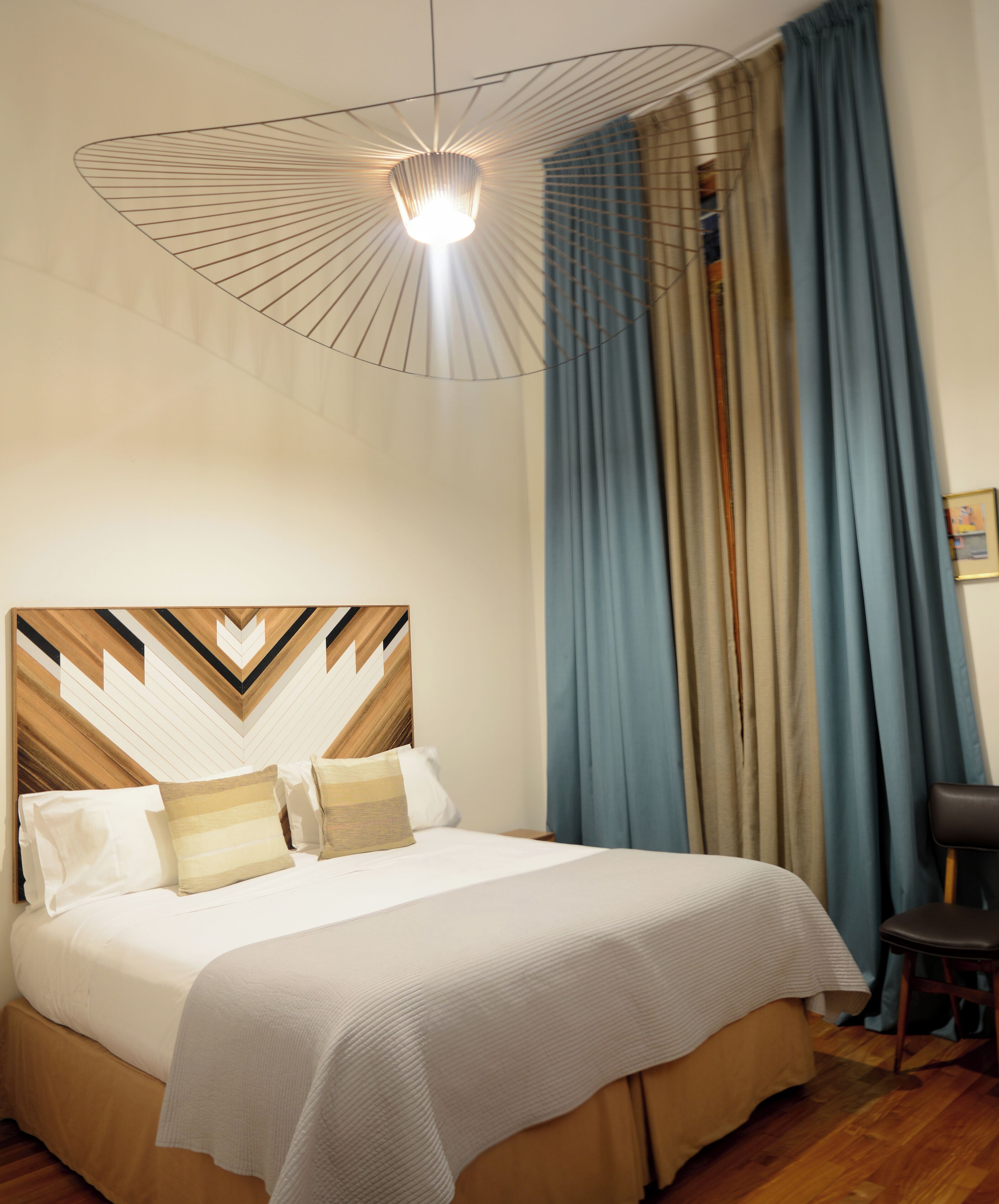 A neatly made bed with a geometric-patterned headboard, a hanging light fixture, and a view of a window with curtains.