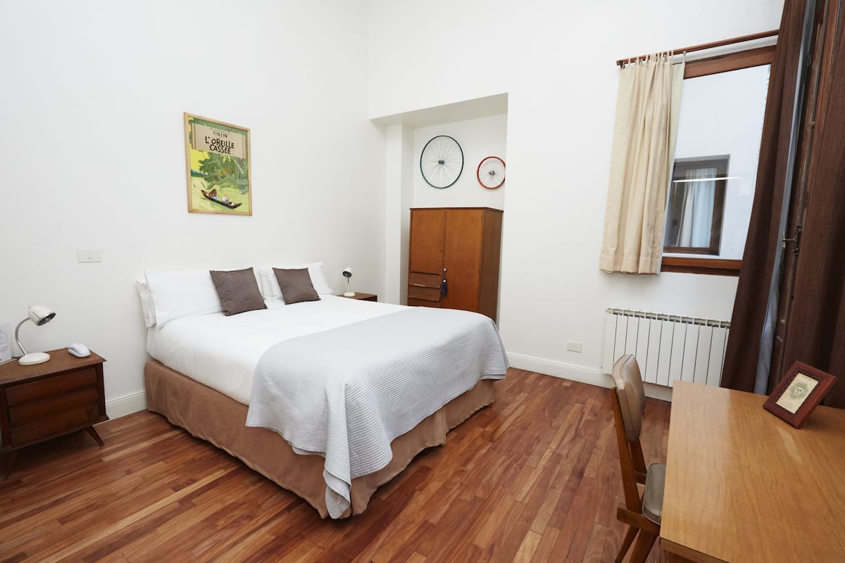 A bedroom with a bed, wooden floor, a wardrobe, a nightstand, a table, and a window with curtains.