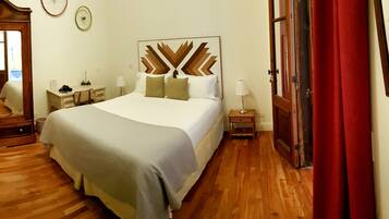 Quarto de hotel com uma cama grande, uma escrivaninha de madeira, uma cadeira, um espelho e um guarda-roupa.